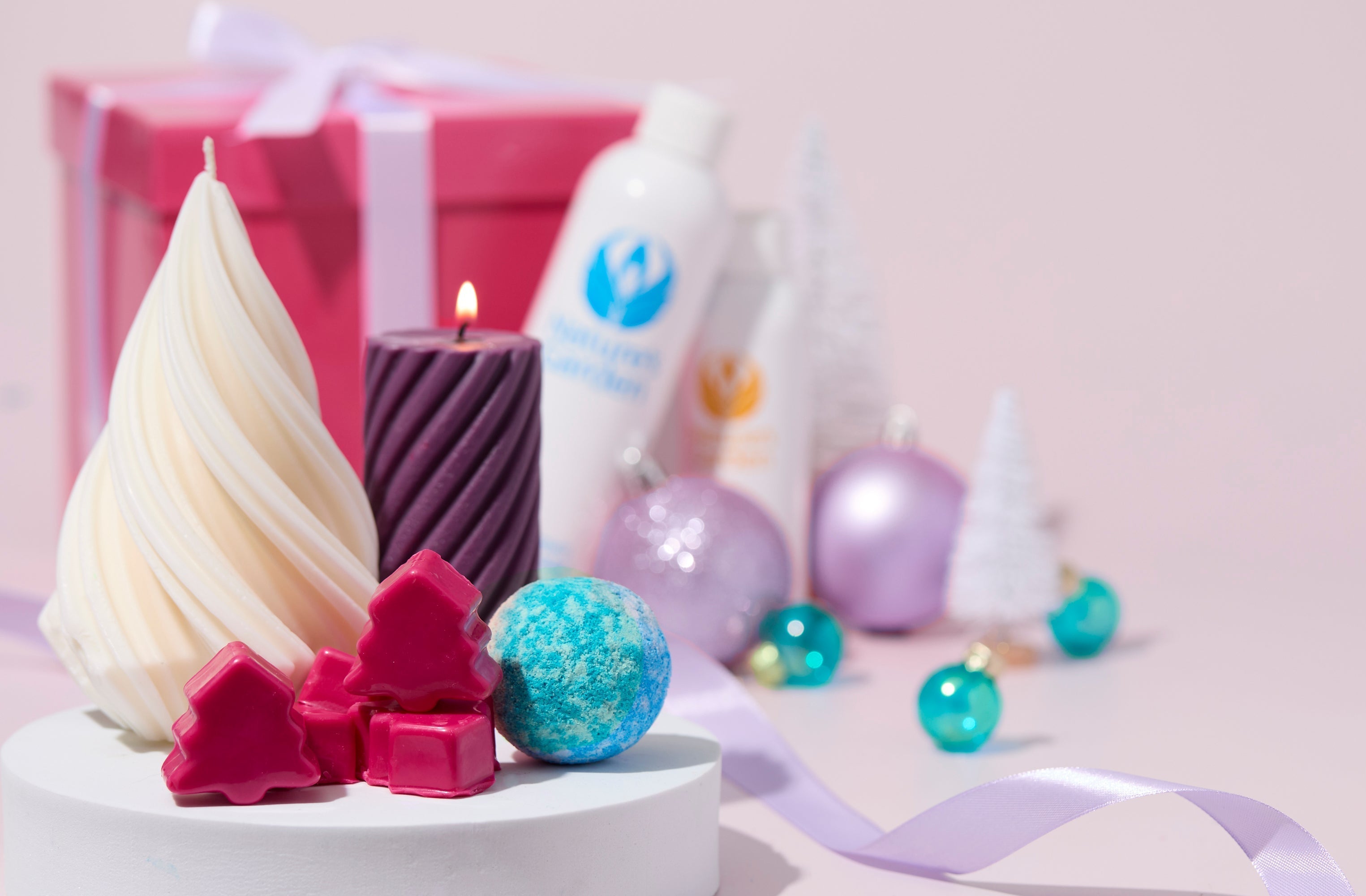 Holiday display with candles, pink tree-shaped melts, and a blue bath bomb on a white pedestal. Nature’s Garden bottles, pastel ornaments, and a pink gift box with a white ribbon sit in the background against a soft pink backdrop.