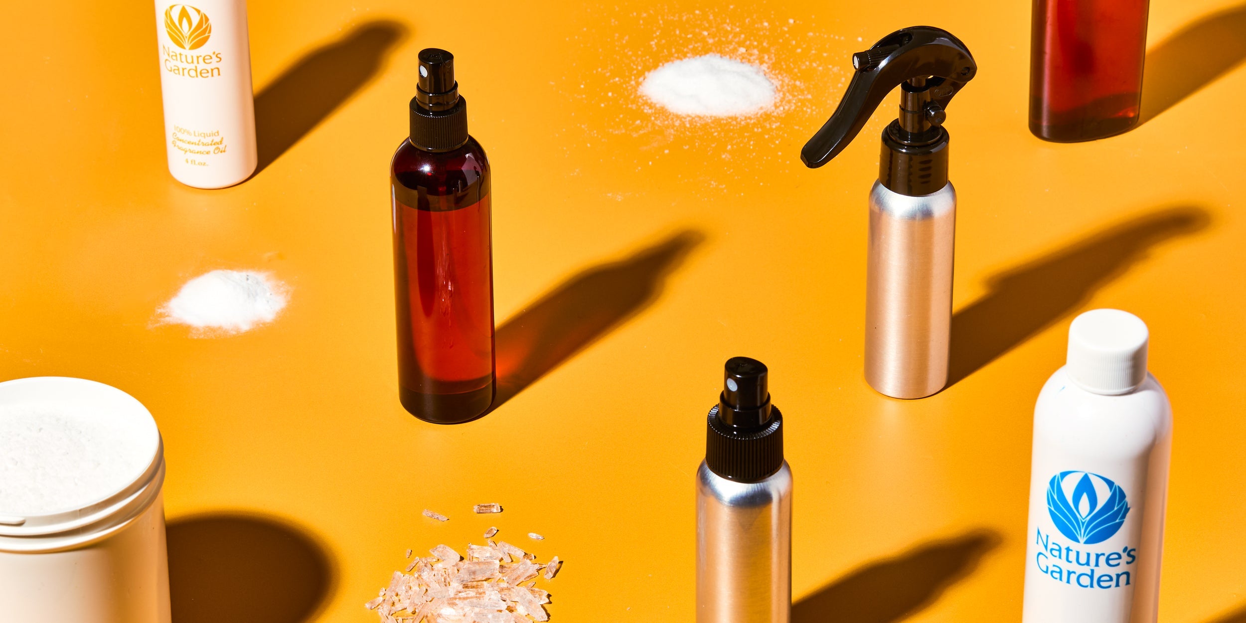 DIY home scent supplies on orange background, including amber and silver spray bottles, Nature’s Garden fragrance oils, powdered ingredients, and plastic jars of white base.