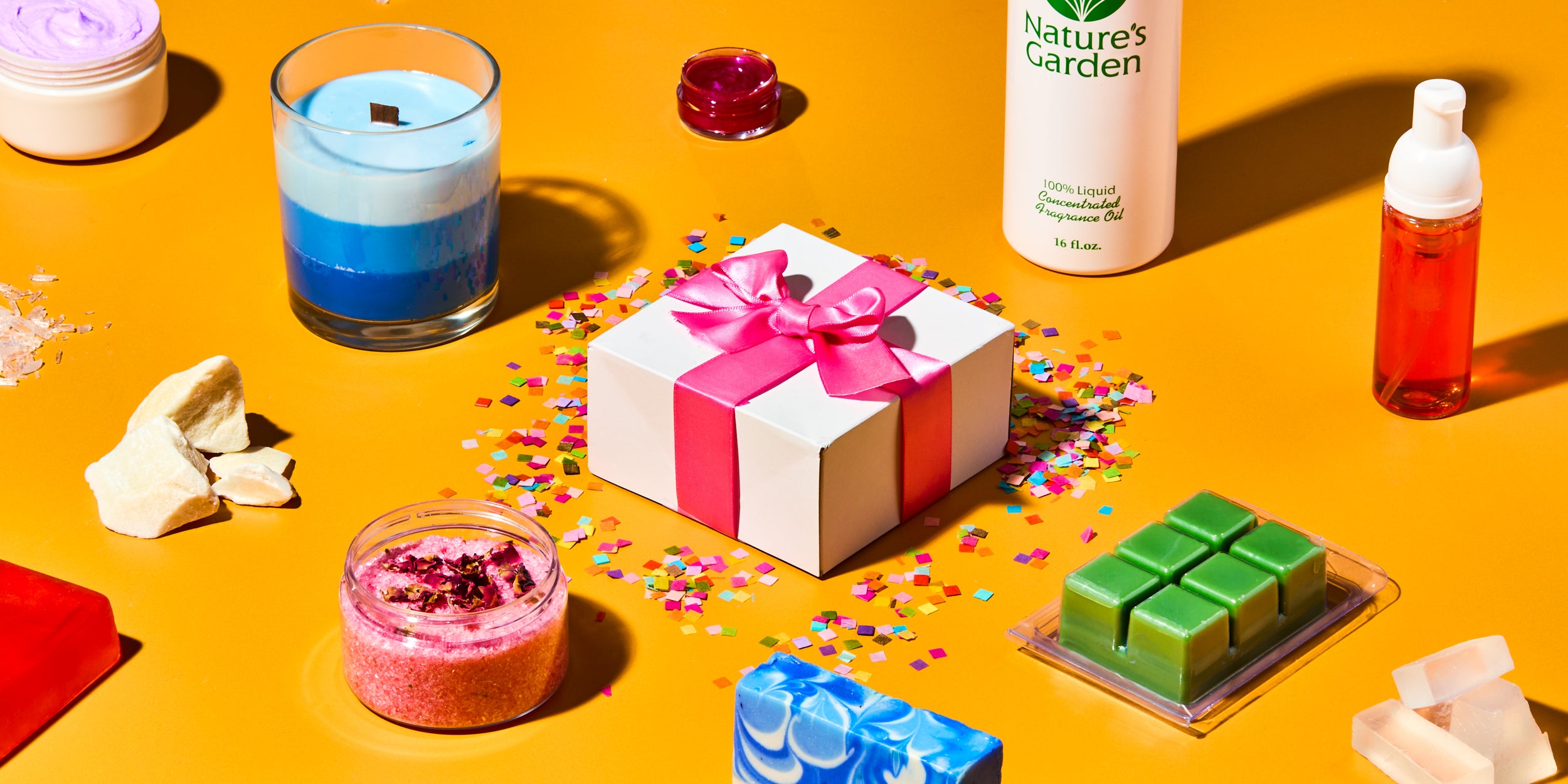 DIY craft supplies and gift box on orange background, featuring Nature’s Garden fragrance oil, layered blue candle, red soap bar, pink whipped body butter, bath salts with dried flowers, wax melts, solid butter chunks, and confetti around a white box with pink bow.