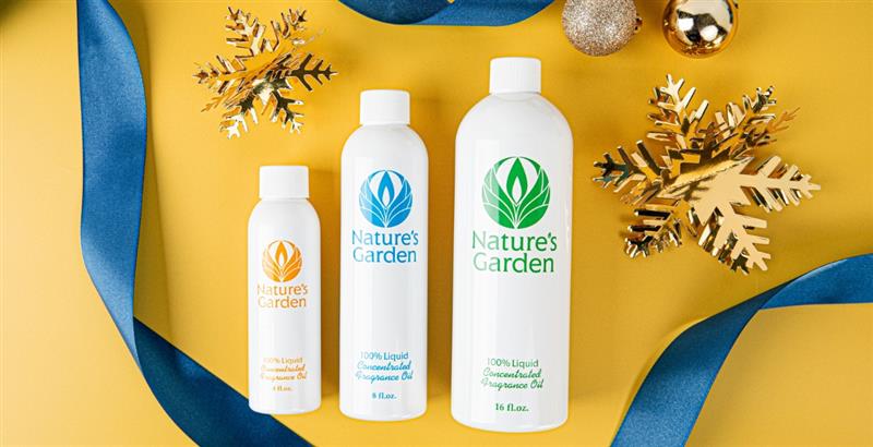 Four Nature’s Garden fragrance oil bottles arranged on a yellow background with gold snowflake ornaments and metallic baubles. A deep blue ribbon loops around the bottles in a loose heart shape.