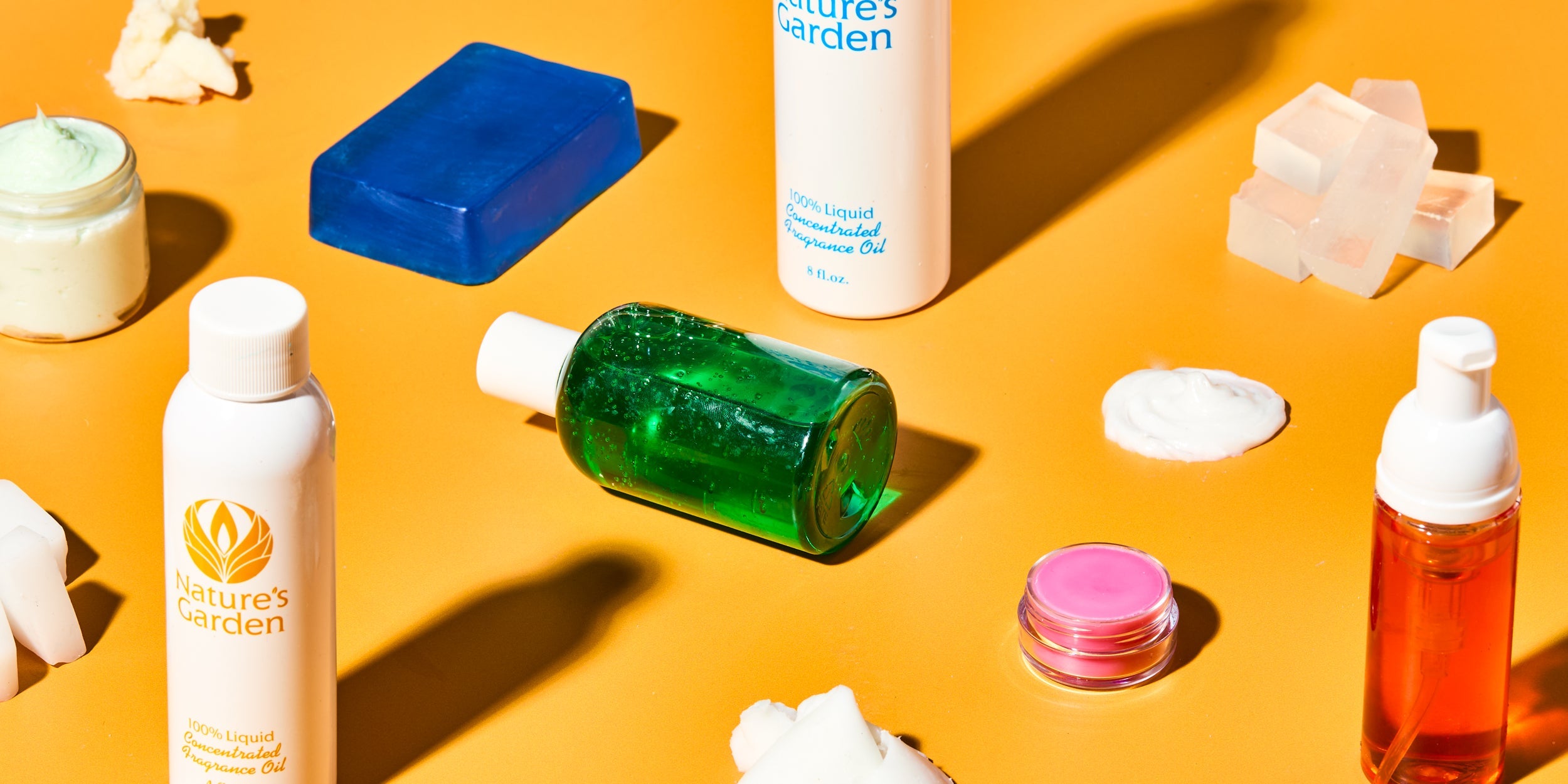 DIY soap and skincare bases on orange background, featuring Nature’s Garden fragrance oils, clear melt and pour soap blocks, blue soap bar, green gel in bottle, foaming wash, whipped cream base, lip balm, and chunks of solid butter.