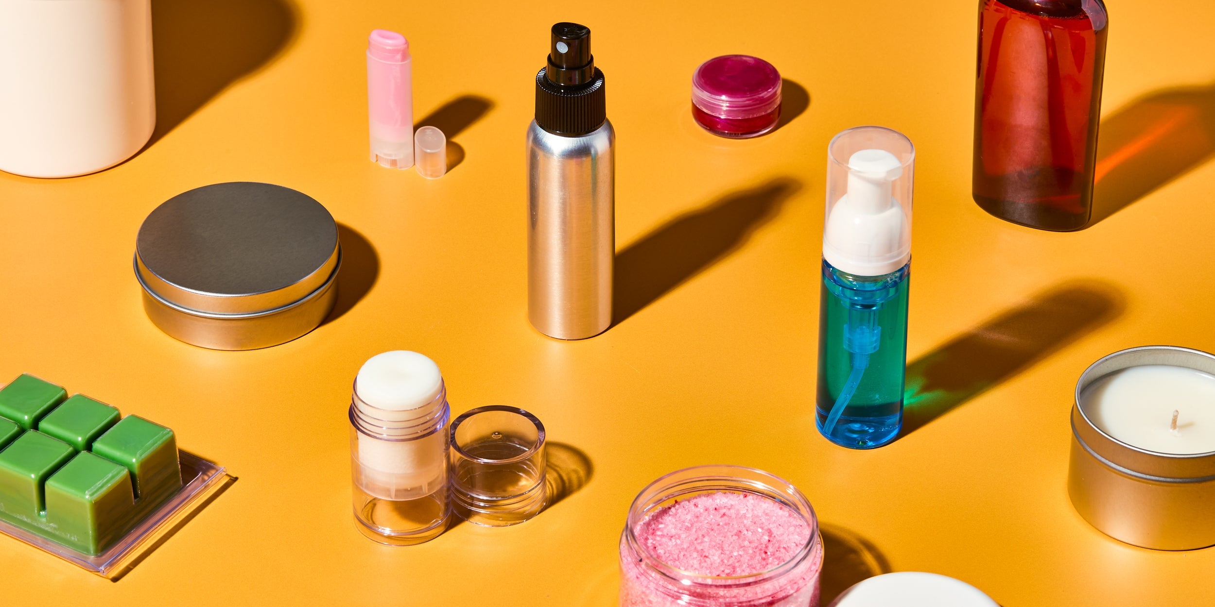 DIY packaging and bath product supplies on orange background, including aluminum spray bottle, foaming pump bottle with blue liquid, lip balm tubes, tins, candle, wax melts, and jars of pink bath salts and lip balm.