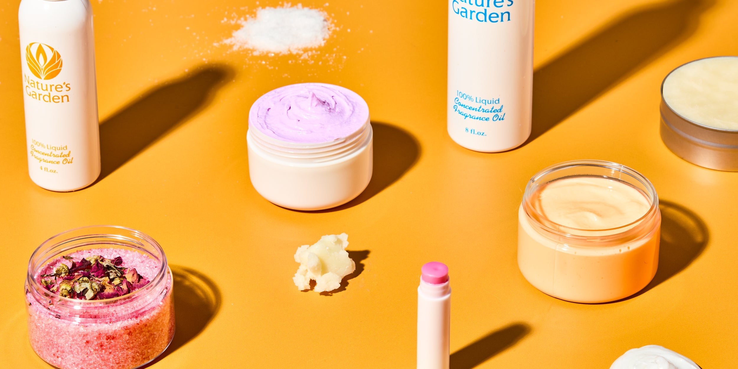 Personal care DIY supplies on orange background, including Nature’s Garden fragrance oil bottles, jars of whipped purple and orange body cream, pink bath salts with dried flowers, lip balm tube, solid butter, and a tin of lotion.