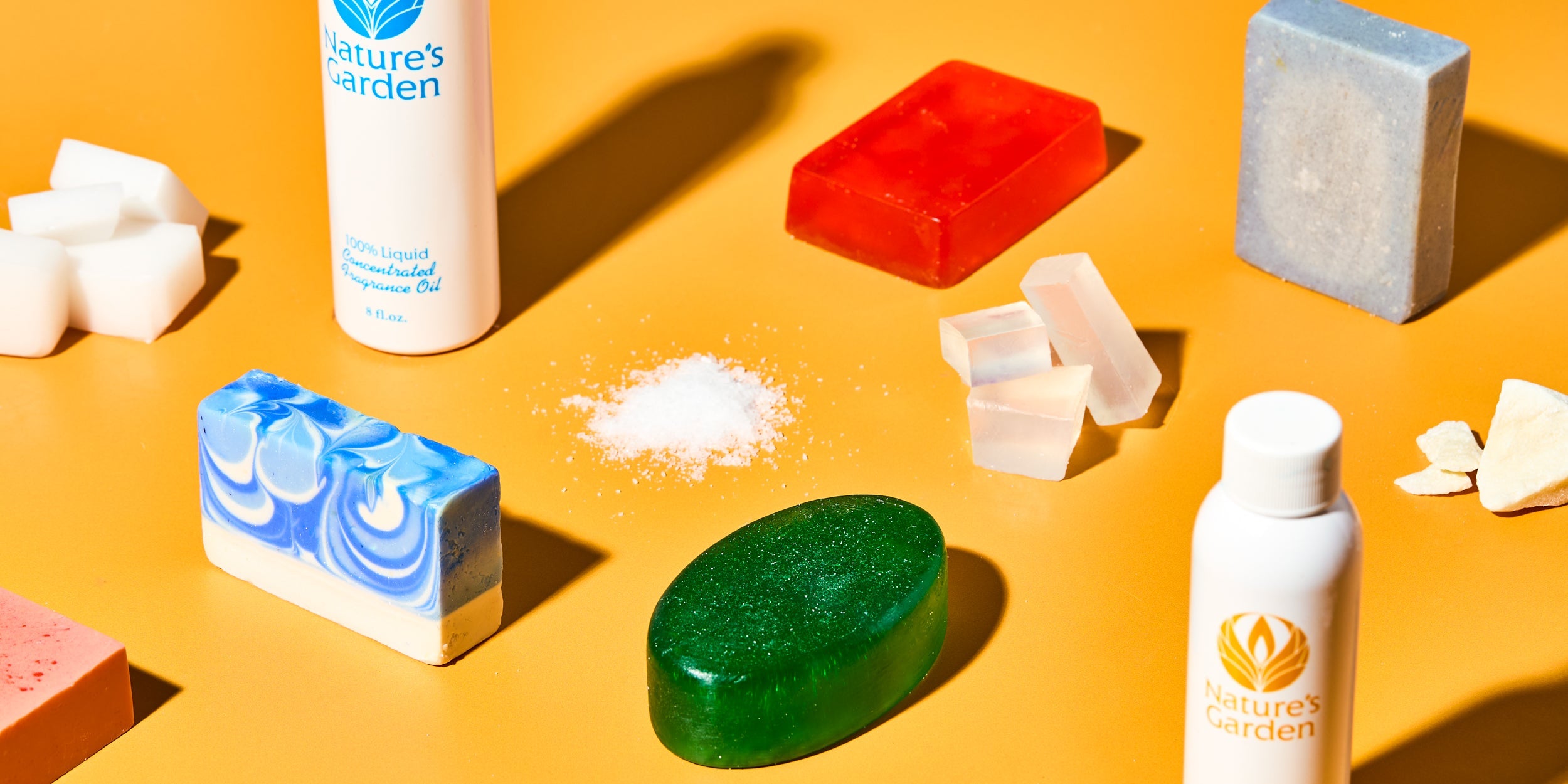Soap making supplies on orange background, featuring Nature’s Garden fragrance oils, colorful soap bars in red, green, blue swirl, and gray, clear melt and pour soap blocks, solid butter chunks, and powdered ingredients.