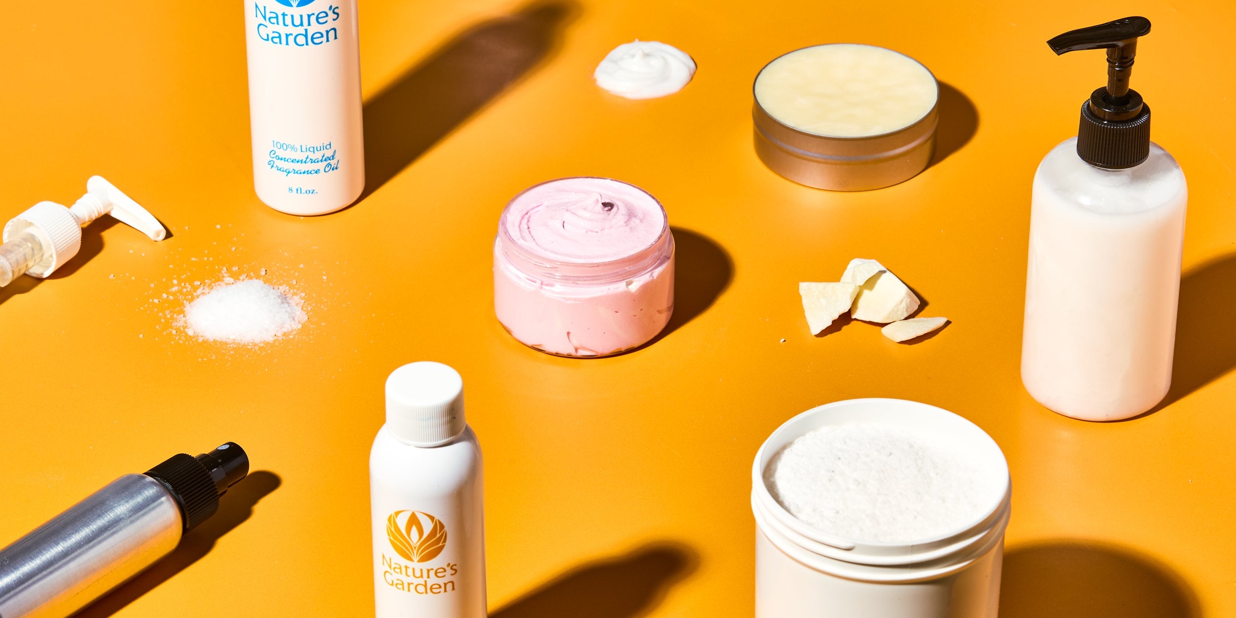 DIY bath and body supplies on orange background, including Nature’s Garden fragrance oil bottles, whipped pink body butter, lotion in pump bottles, powdered ingredients, solid butter pieces, and a tin of balm.