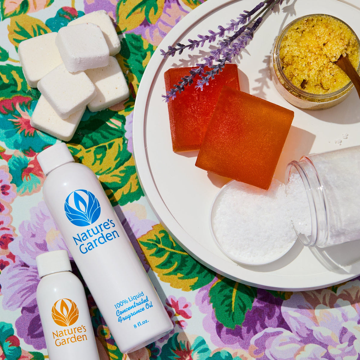 Flat lay of handmade soap supplies on floral fabric: white soap cubes, orange bars on a plate with lavender sprigs, jars of bath salts and sugar scrub, and bottles of fragrance oil.