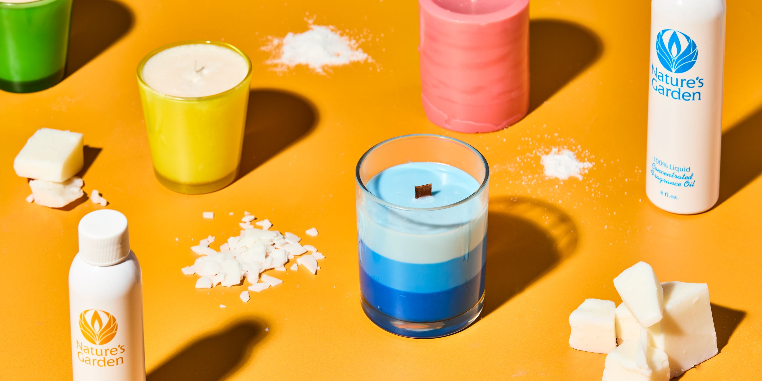 DIY candle making supplies on orange background, featuring layered blue candle in glass, yellow and pink candles, Nature’s Garden fragrance oils, soy wax flakes, wax chunks, and powdered ingredients.