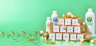 Festive holiday scene on a green background featuring a Nature’s Garden advent-style display with numbered boxes (1–12), surrounded by gold ribbon, confetti, and ornaments, with three Nature’s Garden fragrance oil bottles placed around the centerpiece.