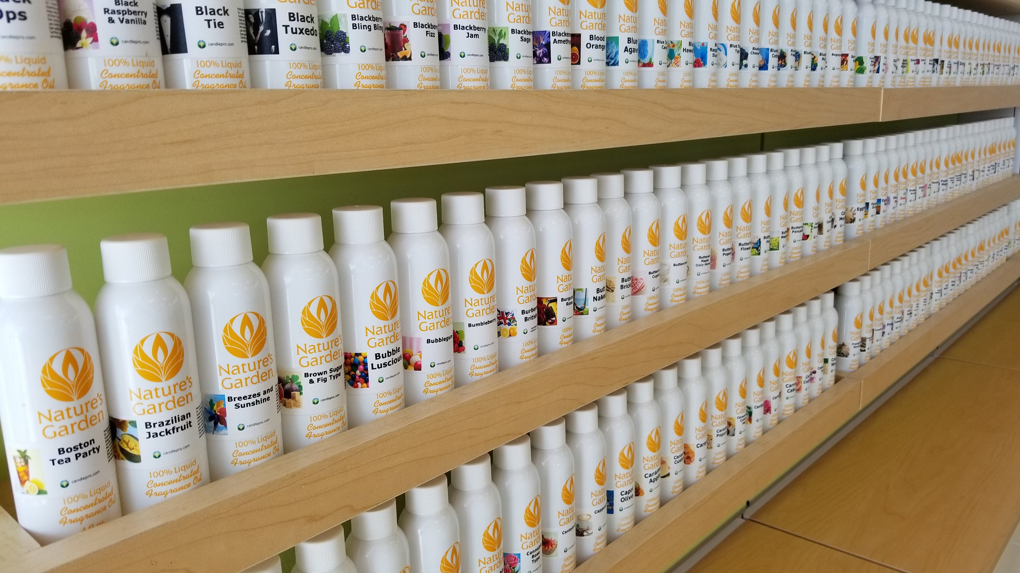 Shelves lined with rows of Nature’s Garden fragrance oil bottles, neatly arranged in white containers with colorful scent labels, displayed on wooden shelving in a bright retail or warehouse setting.