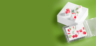 Wax melt clamshells and cubes styled on a bright green background. The melts are white with embedded red and green chunks, giving a festive, Christmas-inspired look. A clear plastic clamshell container sits open in the foreground, while several individual cube melts are displayed on a white block in the background.