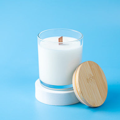 White candle in a glass container with a wooden wick and lid on a blue background