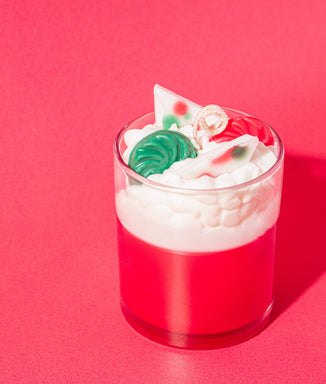 Festive red and white candle in a glass jar topped with whipped wax and decorative embeds shaped like candy pieces and peppermint bark, set against a matching red background.
