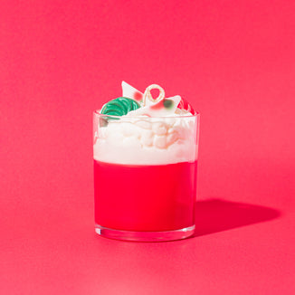 A red and white layered candle in a clear glass jar, topped with whipped wax and wax embeds shaped like holiday candies in red, green, and white, set against a bright pink background.
