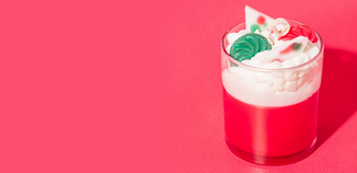 Red and white layered candle in a clear glass, topped with whipped wax and festive wax embeds resembling candies, set against a bright pink background.