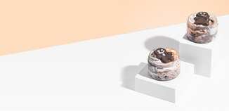 Two small clear jars filled with swirled wax melt “whipped” topping and cookie-like gingerbread embeds sit on white display blocks against a peach and white background.