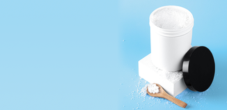 Open white container of powdered laundry detergent with some powder spilled onto the surface and a small wooden scoop beside it, set against a light blue background.