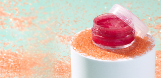 Small clear jar of bright pink lip scrub sitting on a white pedestal, surrounded by scattered peach-colored sugar crystals on a pastel aqua background.
