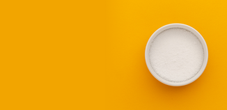 Top-down view of a white bowl filled with fine white lye powder on a bright yellow background.