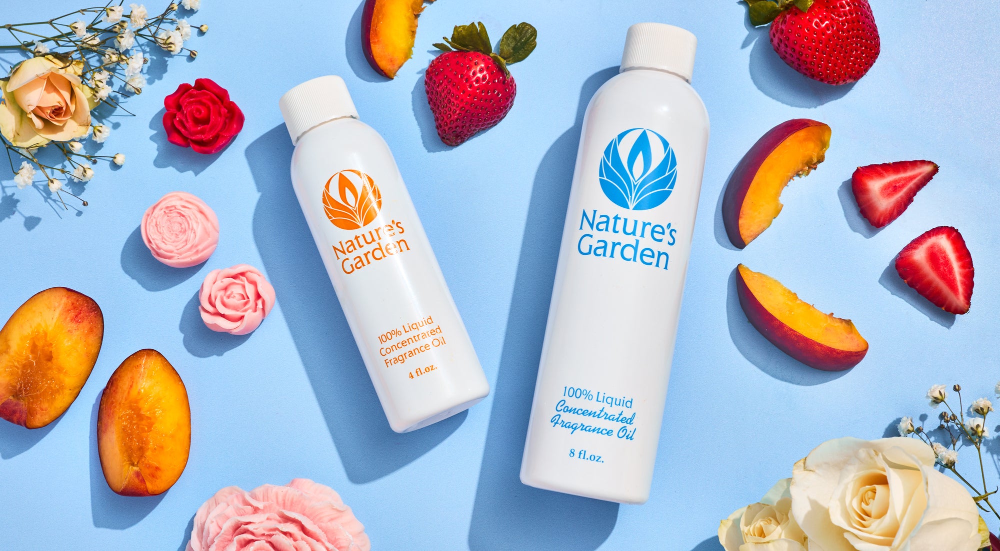 Flat lay of two Nature’s Garden fragrance oil bottles on a blue background, surrounded by strawberries, peach slices, and pastel flowers including roses and baby’s breath.