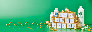 A festive flat lay featuring three bottles of Nature’s Garden fragrance oils beside a 12-day advent calendar made of small gold gift boxes with white numbered labels. The setup is decorated with gold ribbon curls, glitter ornaments, and confetti on a green background.
