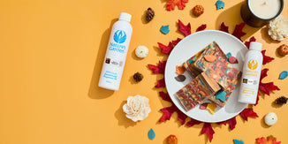 Two bottles of Nature’s Garden concentrated fragrance oil with marbled autumn-colored soap bars on a white plate, surrounded by fall leaves, pinecones, and mini pumpkins on a warm orange background.