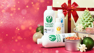 Holiday display featuring Nature’s Garden fragrance oils surrounded by gold and green ornaments, a lit Christmas tree candle, and festive soaps. A large gift wrapped with a red ribbon sits in the background against a sparkling red backdrop.