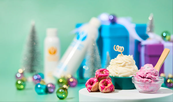 Whipped cupcake candle and pink glazed donut melts displayed on white pedestals with colorful ornaments, mini silver trees, and blue gift boxes in the background. The setup has a festive holiday feel with soft green tones and playful pops of color.