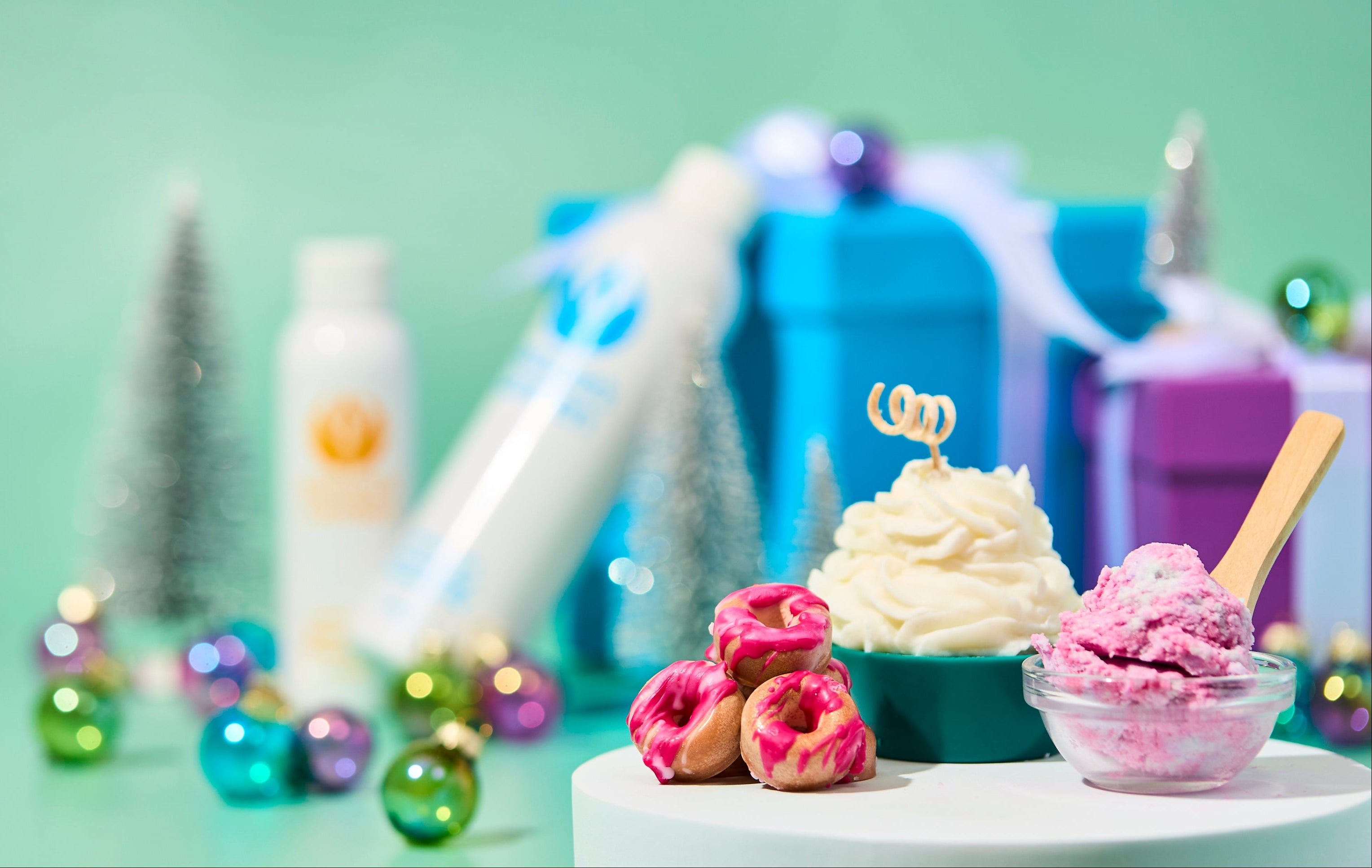 Holiday scene with whipped cupcake candle, pink bath treats, and a small scoop of scrub on white pedestals. Colorful ornaments, silver trees, and blue gift boxes decorate the mint green background for a festive, spa-inspired display.