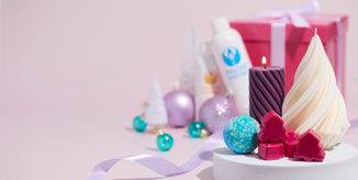Holiday-themed display featuring decorative candles, pink tree-shaped soaps, and a blue bath bomb on a white pedestal. In the background are Nature’s Garden fragrance bottles, pastel ornaments, and a pink gift box with a white ribbon against a soft pink backdrop.