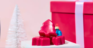 Pink Christmas tree–shaped wax melts arranged on white pedestal stands, surrounded by blue ornaments, a white mini tree, and a large pink gift box with a white ribbon against a soft pink background.
