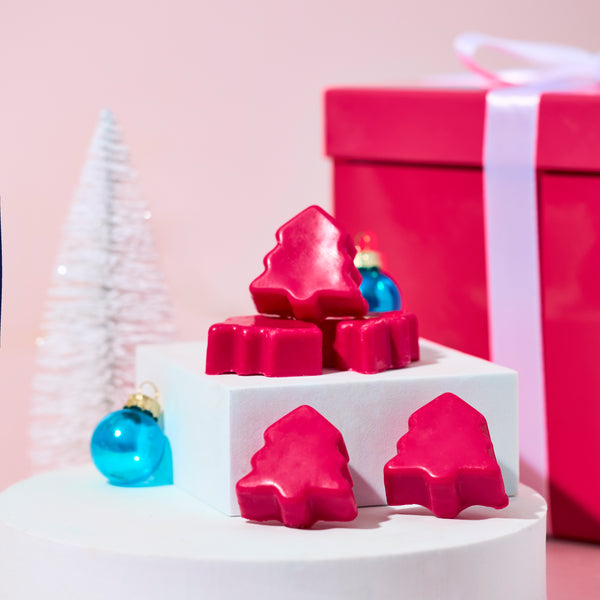 Pink Christmas tree–shaped wax melts arranged on white pedestal stands, surrounded by blue ornaments, a white mini tree, and a large pink gift box with a white ribbon against a soft pink background.
