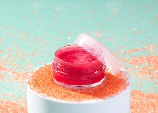 Open jar of shimmering pink lip gloss sitting on a white pedestal dusted with orange glitter, against a mint green background with scattered sparkles.