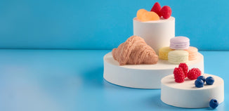 Croissant, pastel macarons, and fresh berries displayed on white pedestal stands against a blue background.