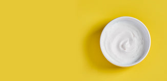 Open jar of white skincare cream with smooth swirl texture on a yellow background.