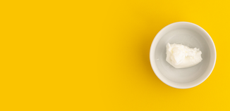 White bowl of creamy lotion on a bright yellow background, photographed from above with minimal styling.