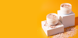 Open jars of pink bath salts resting on white blocks with scattered salt crystals on a bold yellow background.
