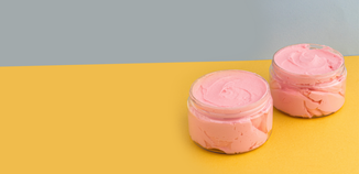 Two small clear jars filled with pink whipped wax sit on a mustard-yellow surface with a gray background, styled with open space for text.