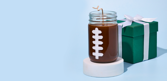 Football-themed jar candle with a curved wick next to a green gift box tied with a white ribbon on a light blue background.