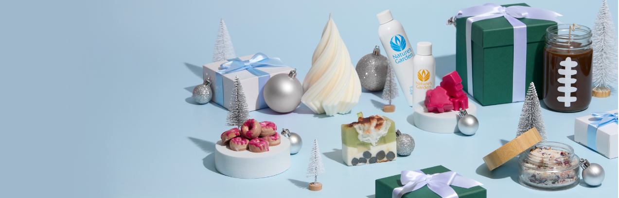 A festive flat lay of handmade bath and body products arranged on a light blue background. The scene includes holiday-inspired soaps, pink wax melts shaped like trees, small jars of bath salts, a candle decorated with a white football design, and bottles labeled “Nature’s Garden.” The products are surrounded by silver ornaments, miniature white Christmas trees, and neatly wrapped green and white gift boxes tied with light blue ribbons, creating a cheerful winter gifting aesthetic.