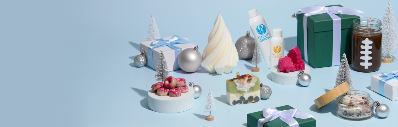 A festive flat lay of handmade bath and body products arranged on a light blue background. The scene includes holiday-inspired soaps, pink wax melts shaped like trees, small jars of bath salts, a candle decorated with a white football design, and bottles labeled “Nature’s Garden.” The products are surrounded by silver ornaments, miniature white Christmas trees, and neatly wrapped green and white gift boxes tied with light blue ribbons, creating a cheerful winter gifting aesthetic.