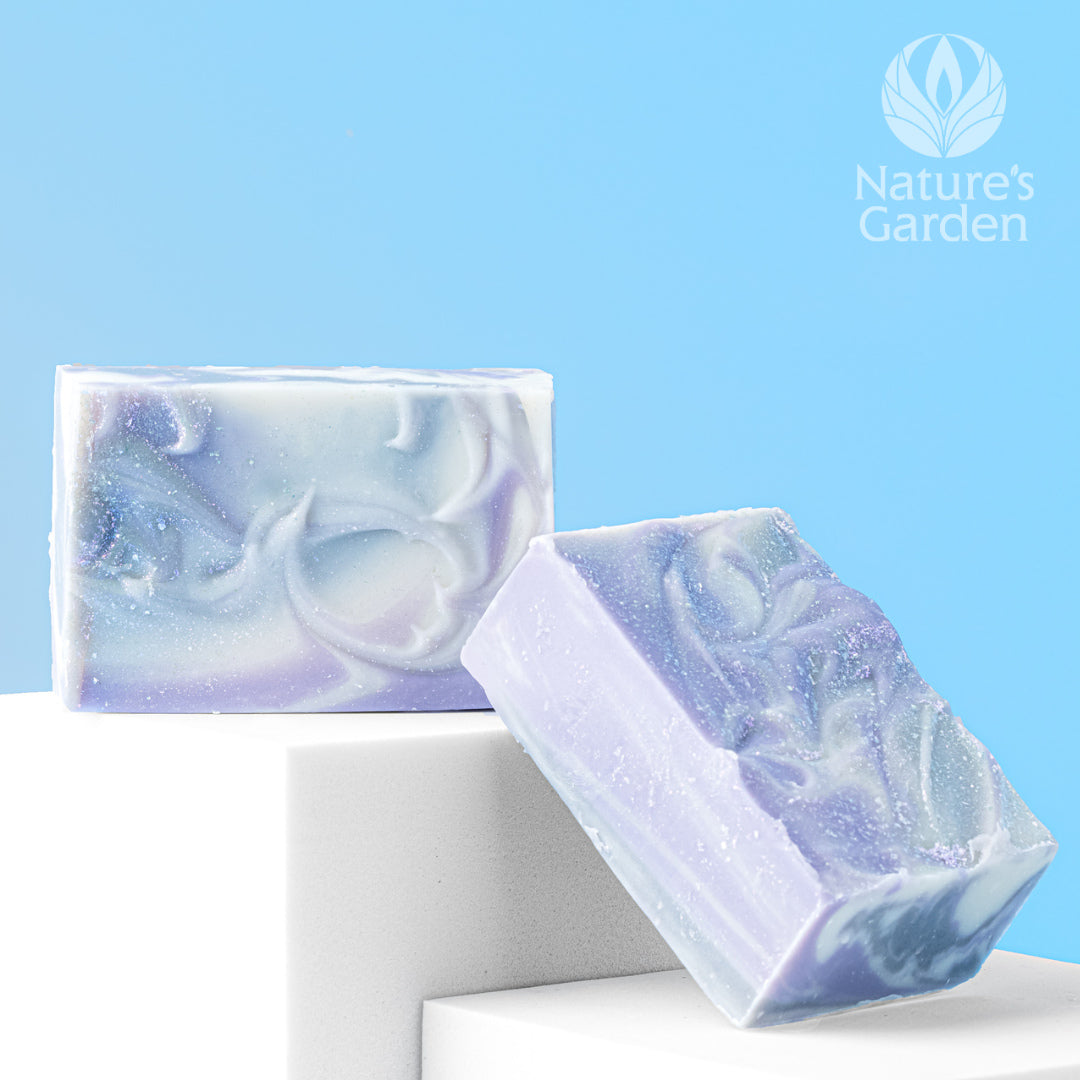Two handcrafted bars of swirled lavender and white cold process soap with a shimmery finish, displayed on white blocks against a light blue background with the Nature’s Garden logo.