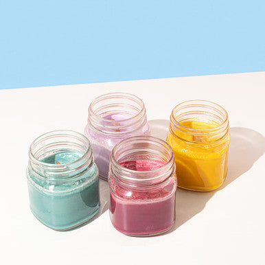 Four multi colored candles in glass jars on a white ground and a blue backdrop
