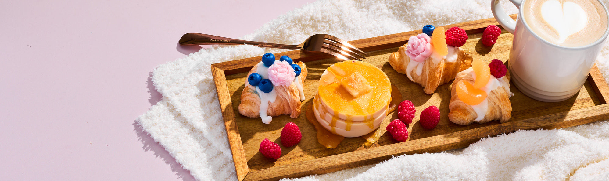 Breakfast-in-bed scene with a wooden tray holding iced mini croissants topped with berries and frosting, a stack of pancakes with syrup, fresh raspberries, and a latte with heart foam on a soft white blanket.