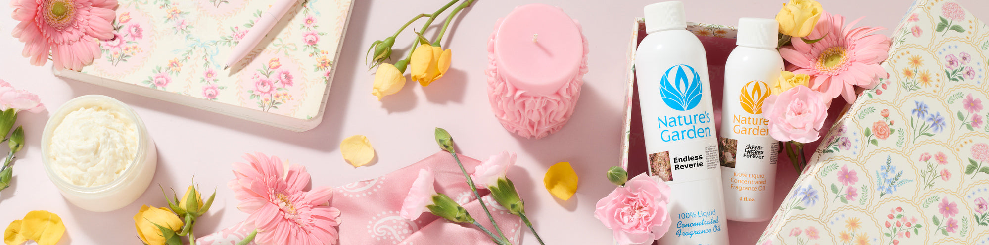 Pink floral flat lay with fragrance oil bottles, a decorative pink candle, whipped body butter, fresh flowers, a floral notebook, and patterned paper arranged on a soft pink background, creating a romantic spring-inspired display.