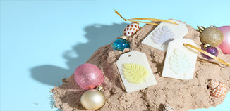 White tag ornaments with pastel tropical leaf designs and gold ribbons on sand, surrounded by colorful bulbs and seashells against a blue background.