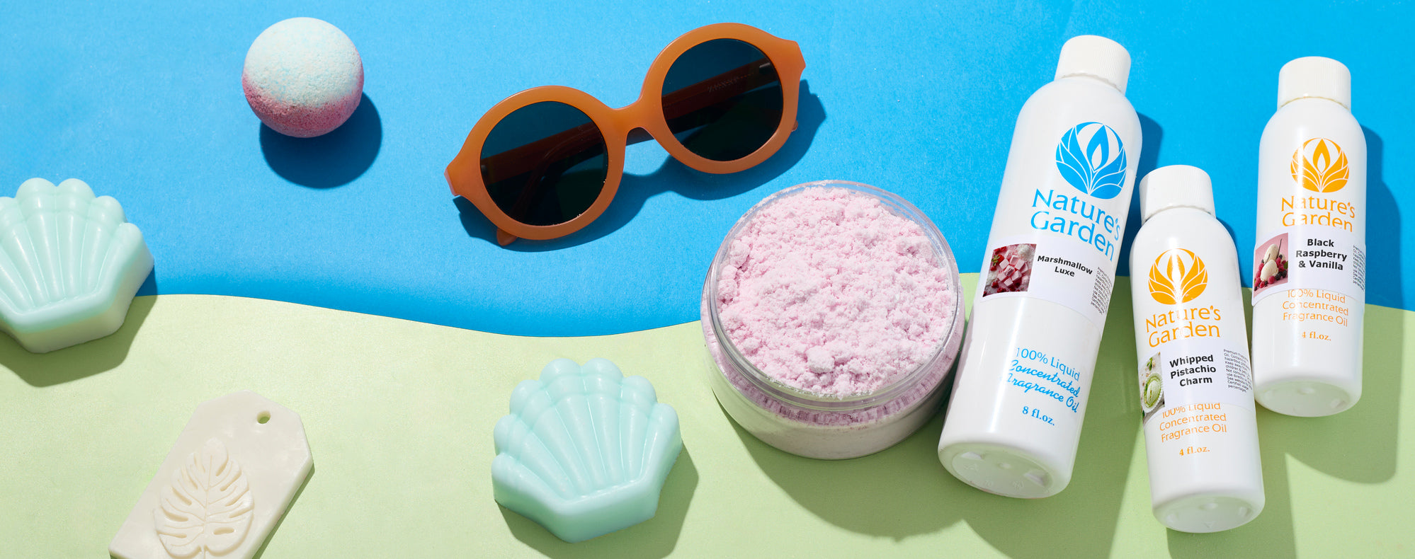 Bright flat lay on blue and green background with round sunglasses, bath bombs, shell-shaped soaps, pink powder in a jar, and bottles of fragrance oil.