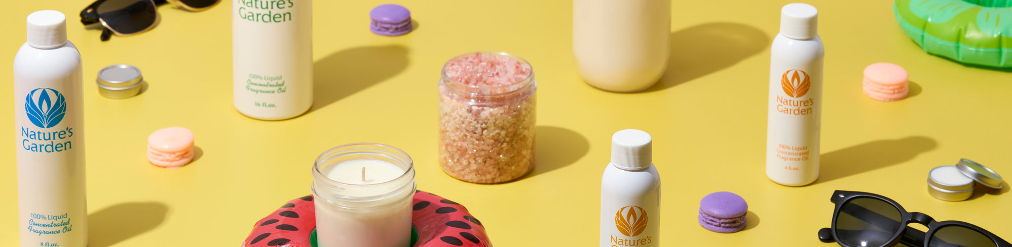 White bottles of fragrance oil, a lit candle in a glass jar, pink bath salts, macarons, sunglasses, and small tins arranged on a bright yellow background with colorful pool floats, creating a playful summer-inspired display.