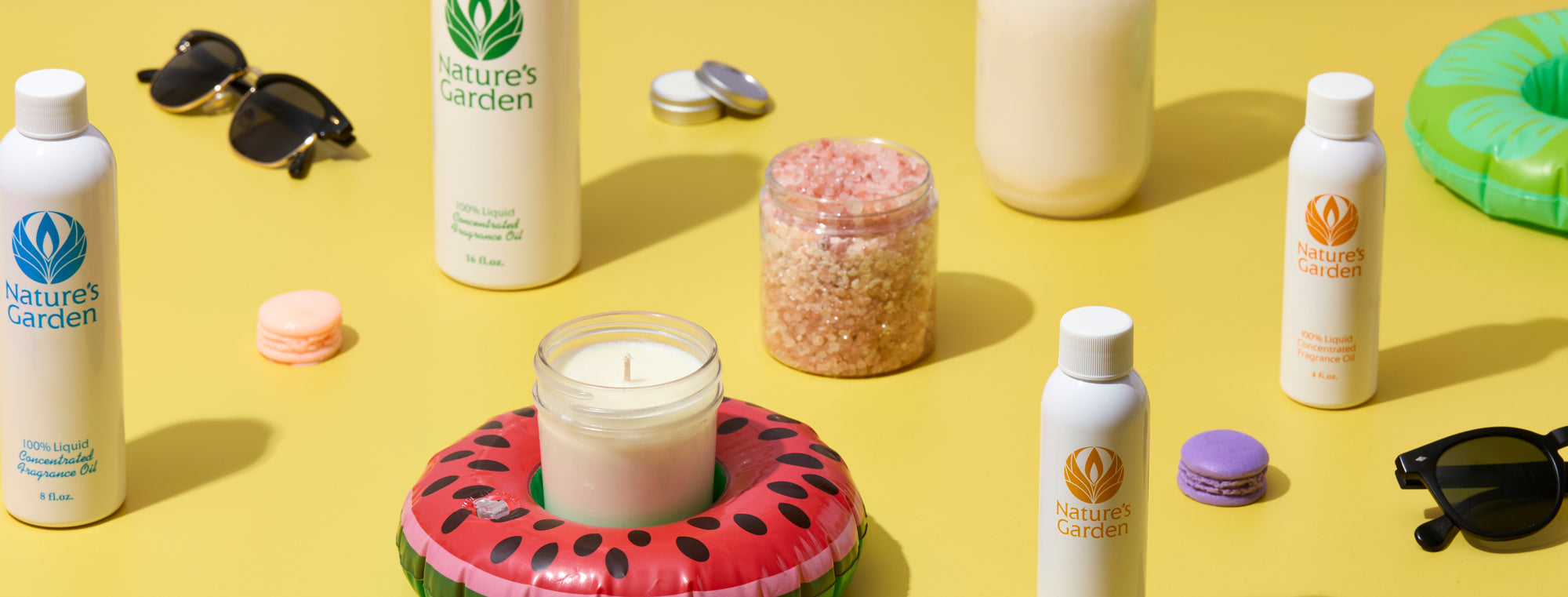 White fragrance oil bottles, a candle in a glass jar on a watermelon float, pink bath salts, macarons, sunglasses, and small tins arranged on a bright yellow background with a green pool float, creating a playful summer-themed display.