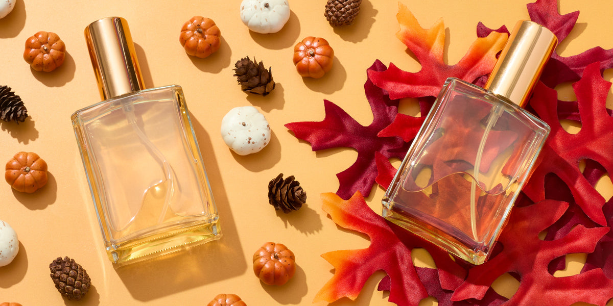 Two clear rectangular perfume spray bottles with gold caps arranged among red autumn leaves, mini pumpkins, white gourds, and pinecones on a warm golden background, creating an elegant fall-themed fragrance display.