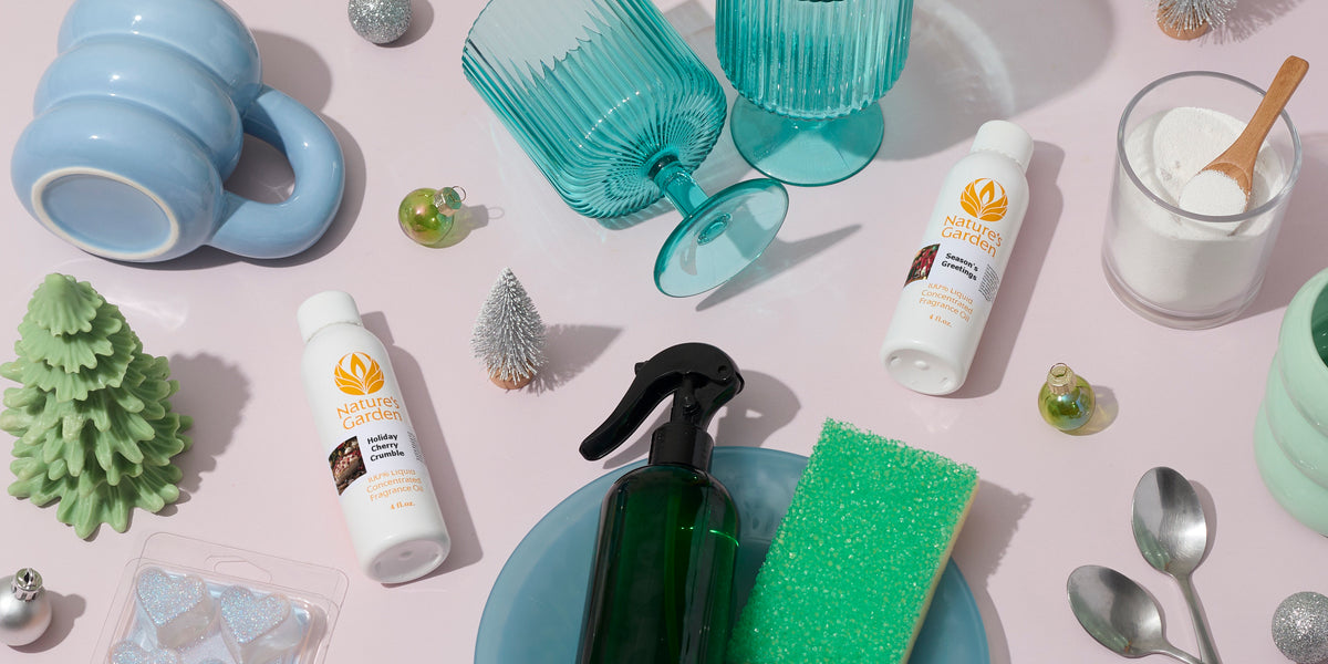 Nature’s Garden holiday fragrances arranged with a green spray bottle, sponge, blue glassware, mugs, ornaments, miniature trees, and cleaning powder on a pale pink surface.